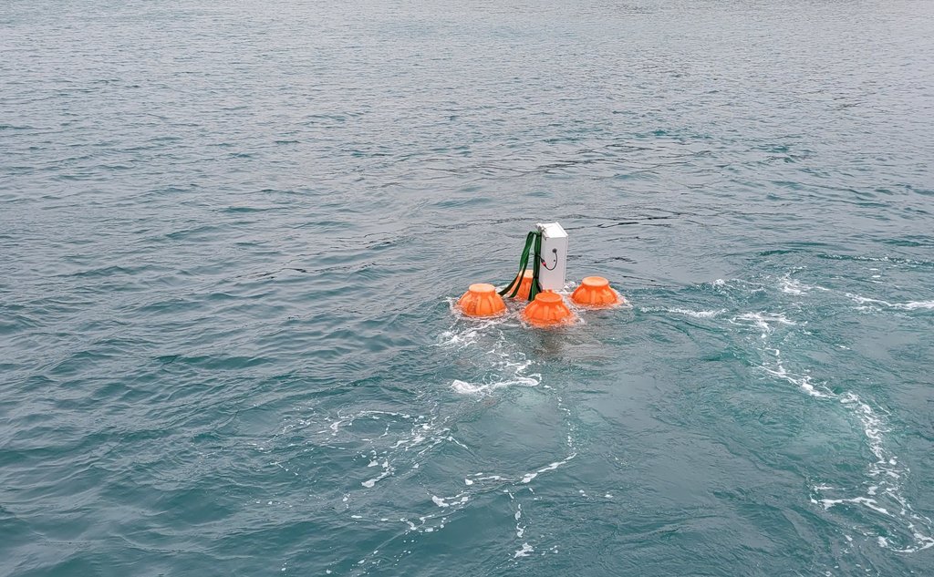 Grå metallboks med oransje flyteelementer på hver side flytende i sjøen.