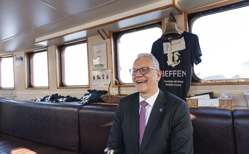 En mann i dress og med briller sitter inne i en liten passasjerbåt.