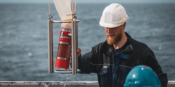 

En mann med kjeledress og hvit hjelm står på dekket av et forskningsfartøy. I bakgrunnen er åpent hav og horisont.