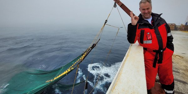 

mann på skip med tommel opp, trål hengende ut i havet bak