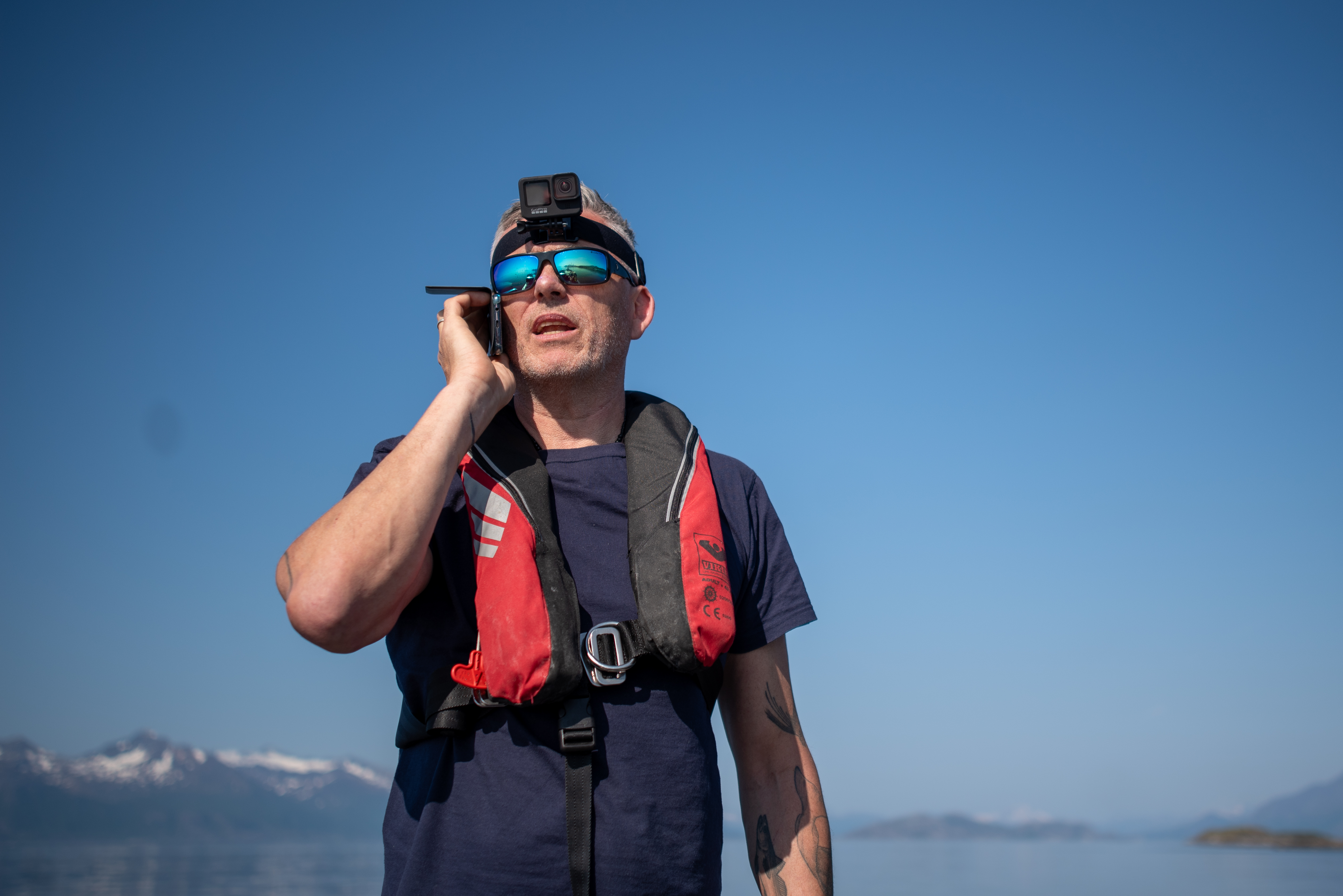Ein mann snakker i telefonen. Han har på seg redningsvest og solbriller. Det er fint vêr og bak han er himmelen heilt blå.