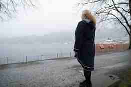 Kvinne i mørk frakk står med ryggen til og ser ut mot sjøen, grått regnvêr.