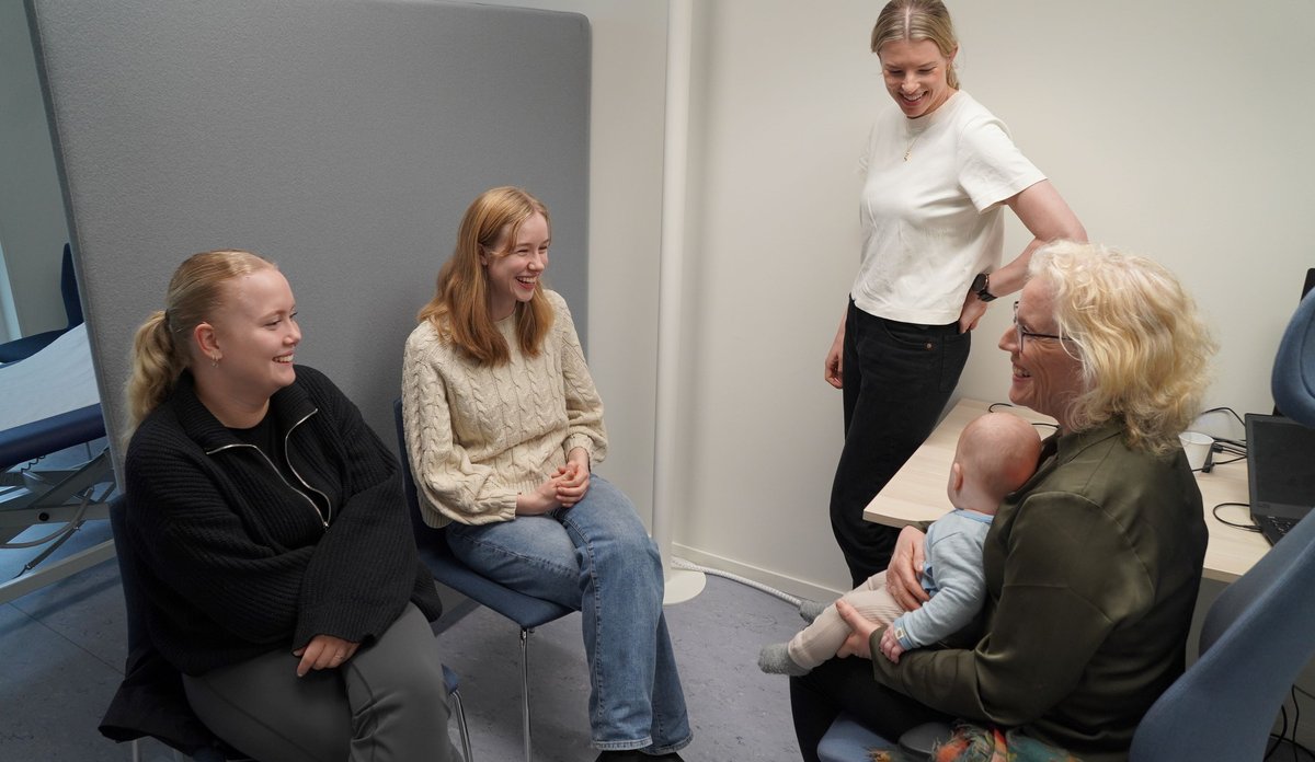 
Fem kvinner på et kontor. To sitter på stol vendt med blikket mot høyre. I midten står en kvinne i hvit t-skjorte. Til høyre sitter en kvinne med et spebarn på fanget.