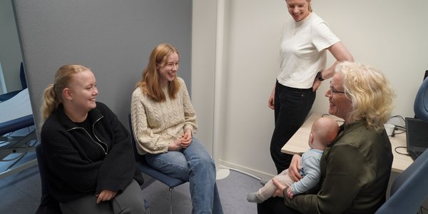

Fem kvinner på et kontor. To sitter på stol vendt med blikket mot høyre. I midten står en kvinne i hvit t-skjorte. Til høyre sitter en kvinne med et spebarn på fanget.
