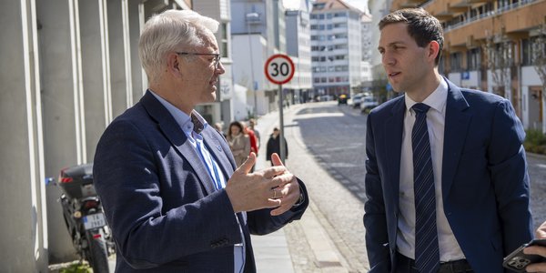 

To dresskledde menn står utenfor et bygg i solskinn