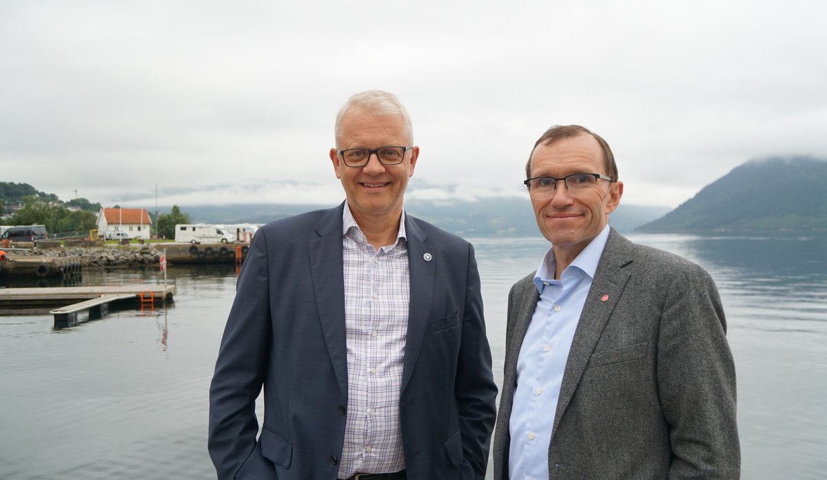 to smilende menn i dress står på en kai med blå fjord i bakgrunnen, i tidlig morgenlys 