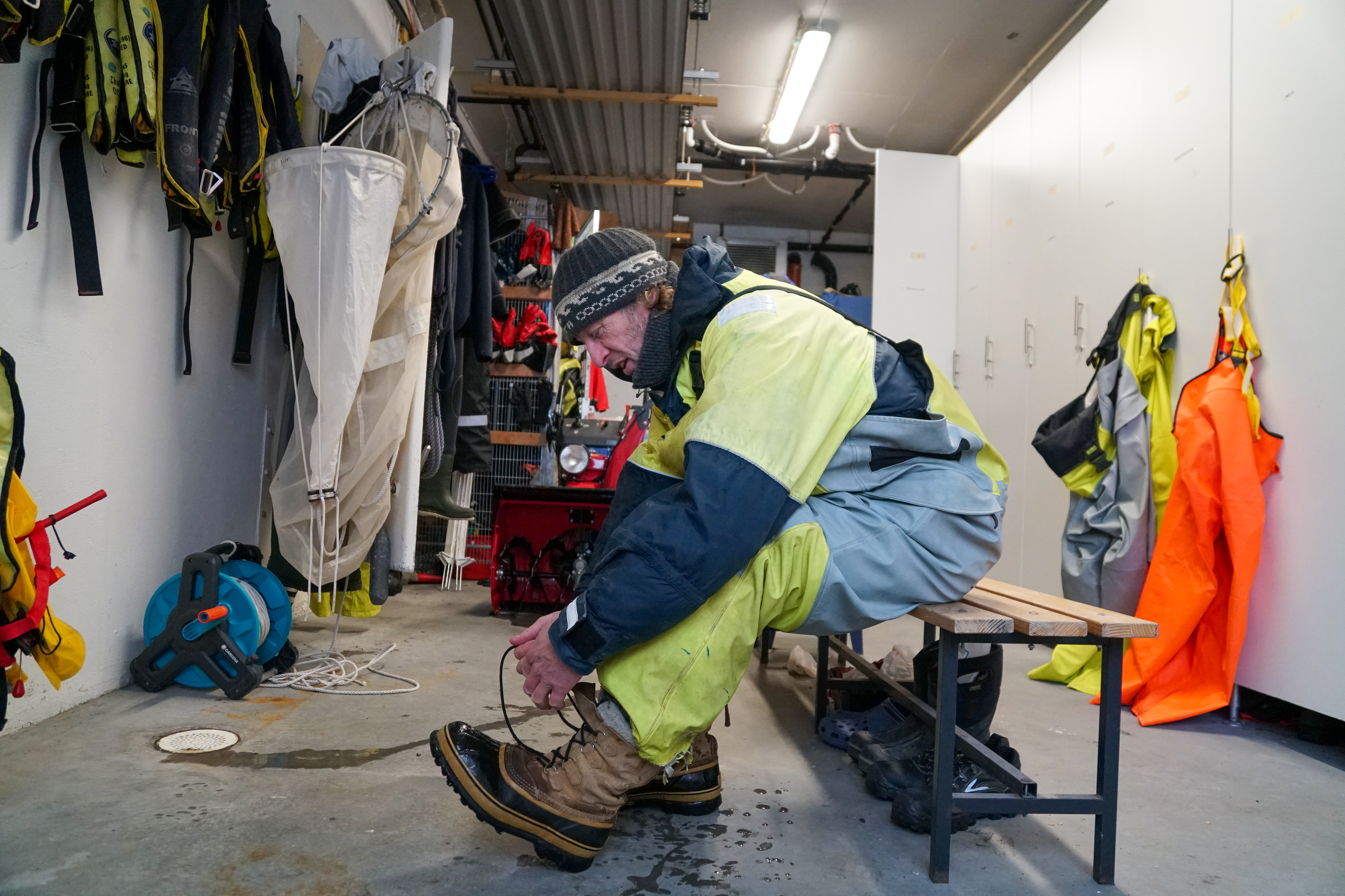 En mann sitter på en benk og knytter skoene sine. Han har på seg arbeidsklær for å vere ute i kulden.