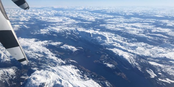 

Fjord omgitt av snøkledde fjell sett ovenfra fra et fly med deler av propell i forgrunnen.