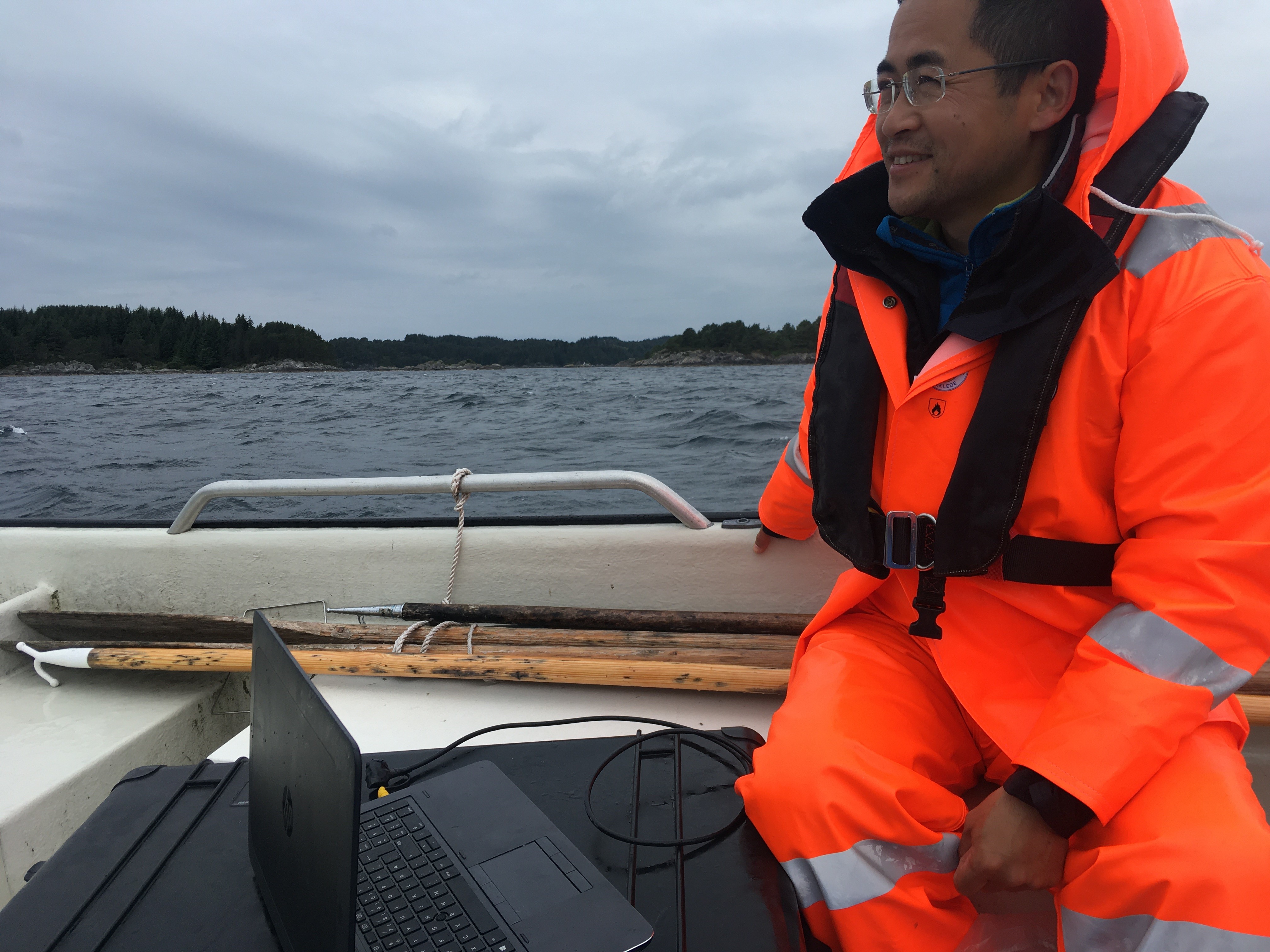 Mann i oransje kjeledress og redningsvest sittende med en bærbar datamaskin i en småbåt ute på fjord med land i bakgrunnen.