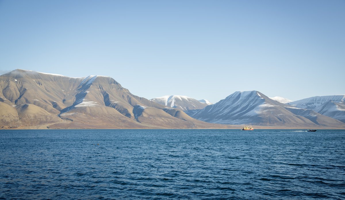 
Nakne, bratte fjell med litt snø på toppen, mørkt, flatt hav og en liten båt 