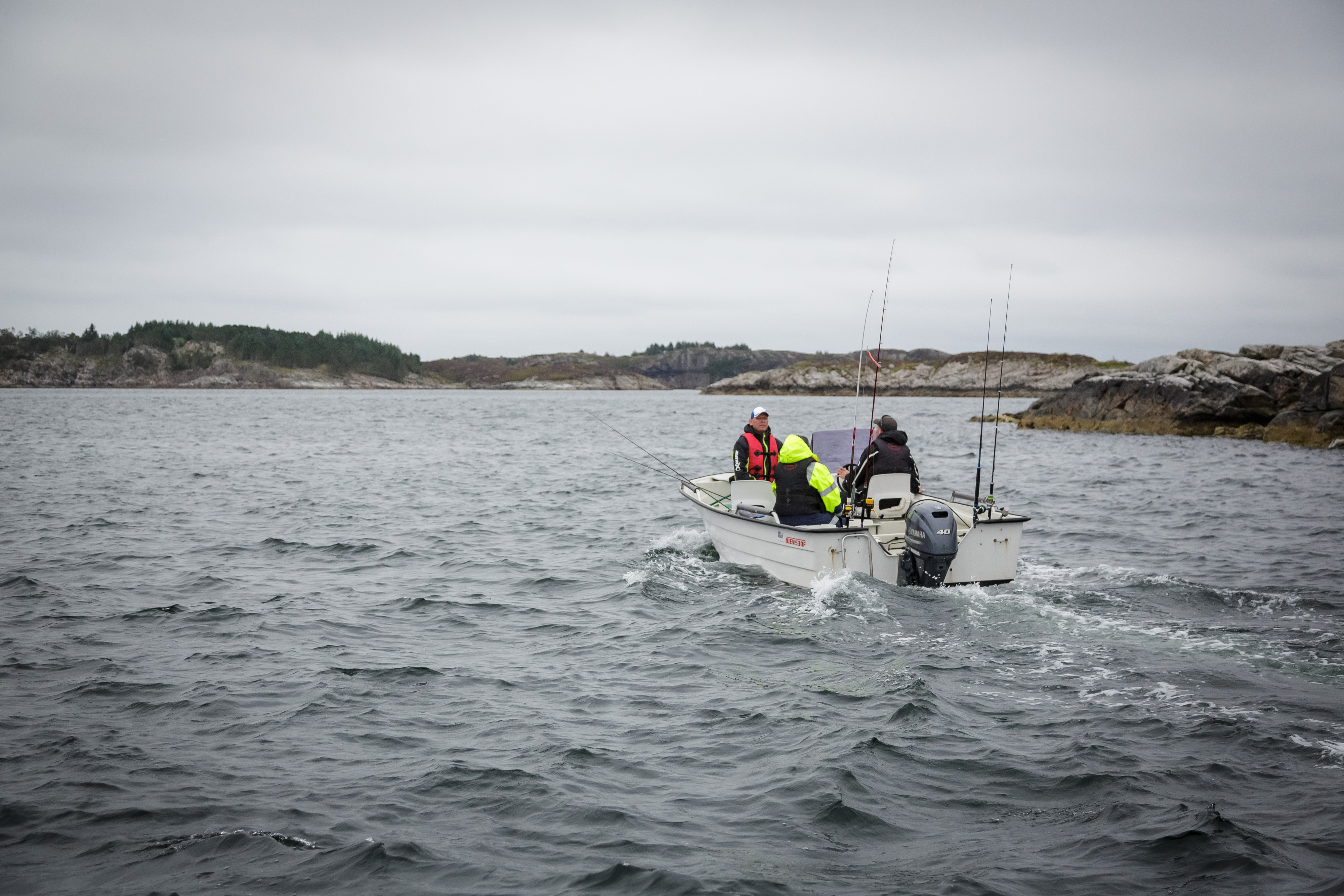 Liten, hvit fritidsbåt med tre personer og flere fiskestenger kjører i kystlandskap.