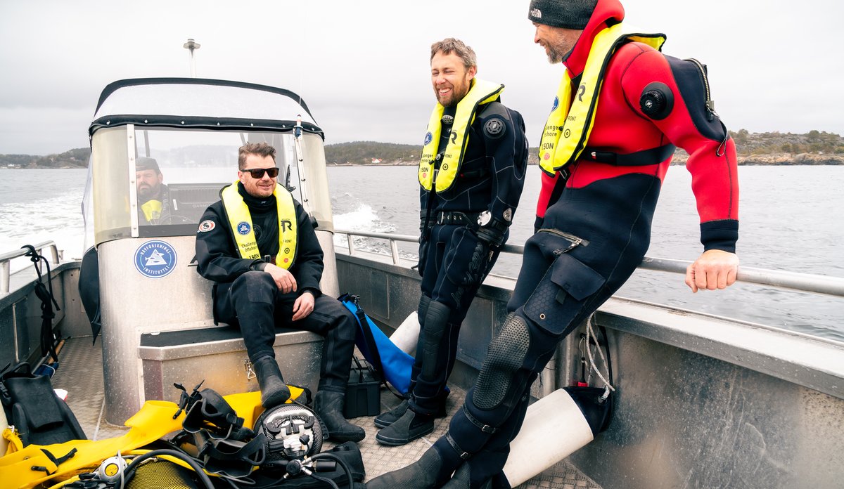 
tre menn sittende i tørrdrakter i en liten småbåt i fart på sjøen, med land i bakgrunnen