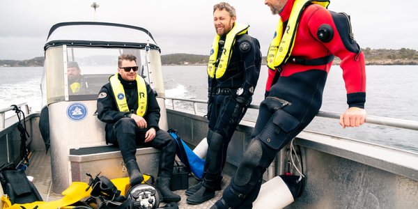 

tre menn sittende i tørrdrakter i en liten småbåt i fart på sjøen, med land i bakgrunnen