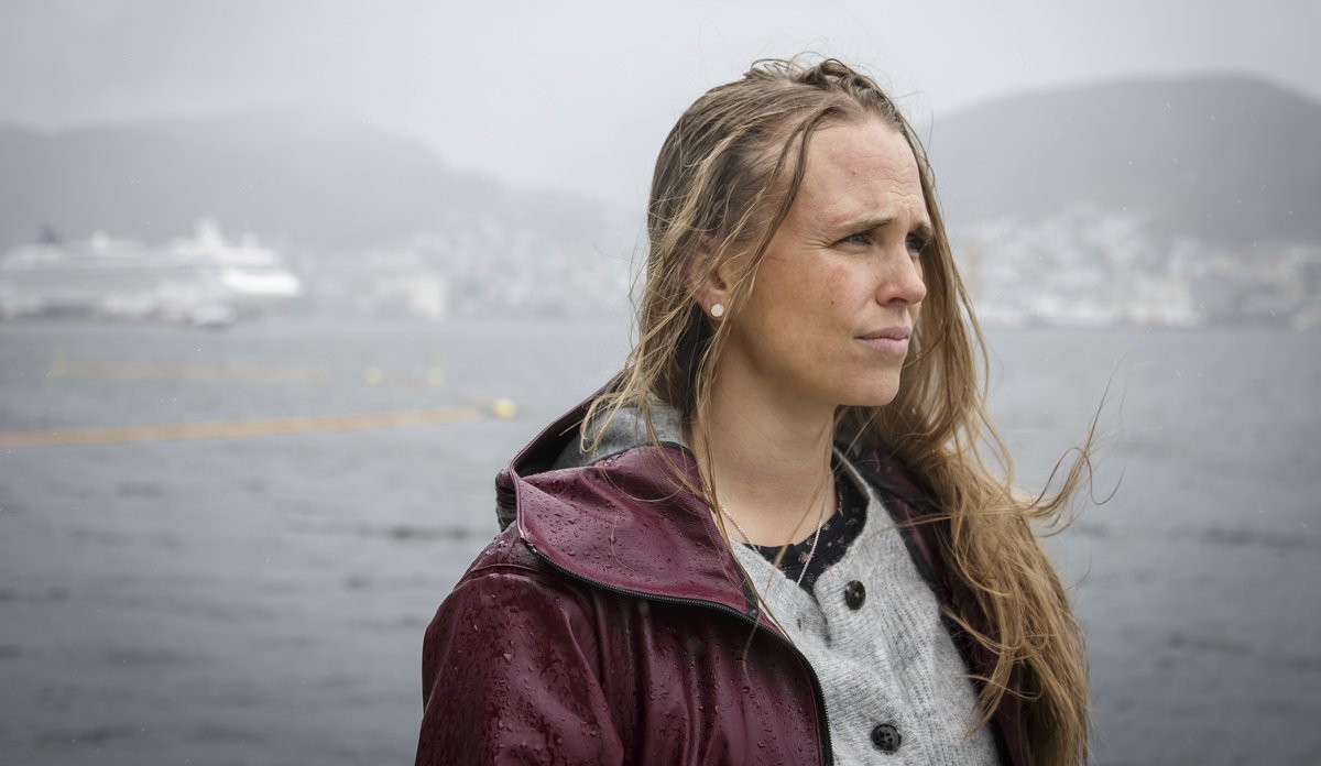
Kvinne med lilla regnjakke og vind og regn i håret, med en fjord, et cruiseskip og bebyggelse i bakgrunnen.