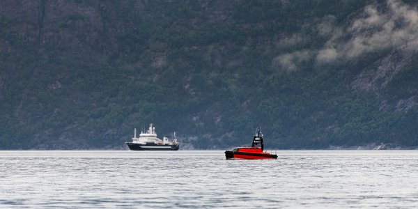 

Foto av en fjord med forskingsfartøy og liten båt-liknende drone i forgrunnen, i en fjord.