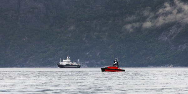 

Foto av en fjord med forskingsfartøy og liten båt-liknende drone i forgrunnen, i en fjord.