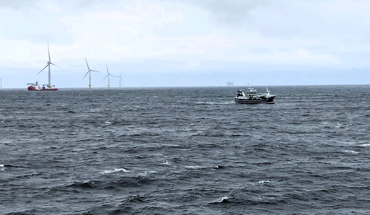 
Fiskebåt passerer vindturbiner i hav med skummende bølger 