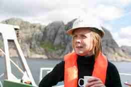 En kvinne med oransje redningsvest og hvit hjelm står på dekk av et fartøy. Hun holder en kaffikopp i hånden og ser ut av bildet. Bak henne ligger Jøssingfjord.