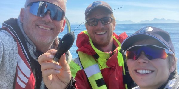 

Tre personar smiler mot kameraet. Dei har alle på seg solbriller og redningsvest. Personen i midten held opp eit lite satellittmerke, som han viser til kamerat.