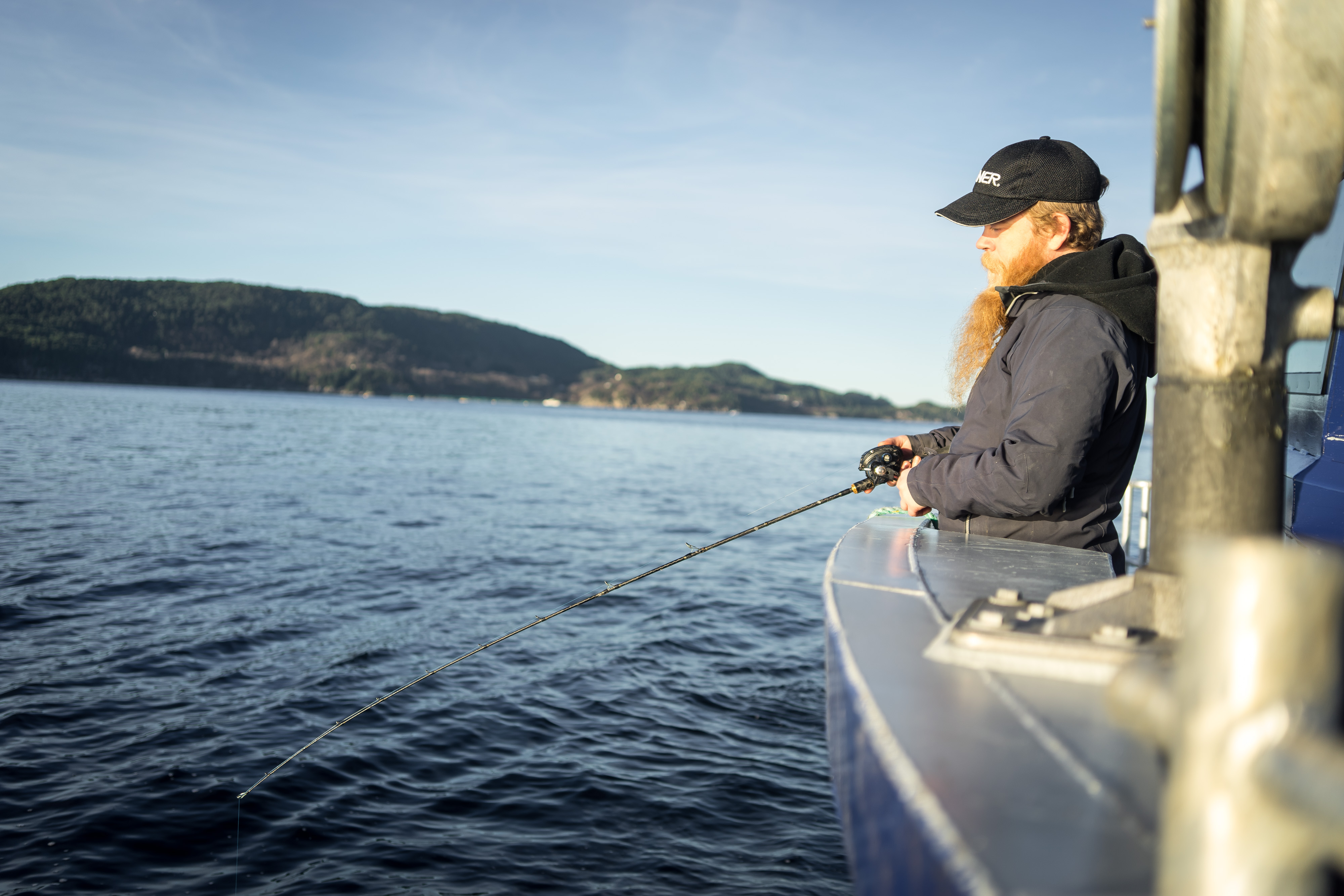 Mann med blå caps og langt skjegg står i båt med fiskestand over rekka og fisker.
