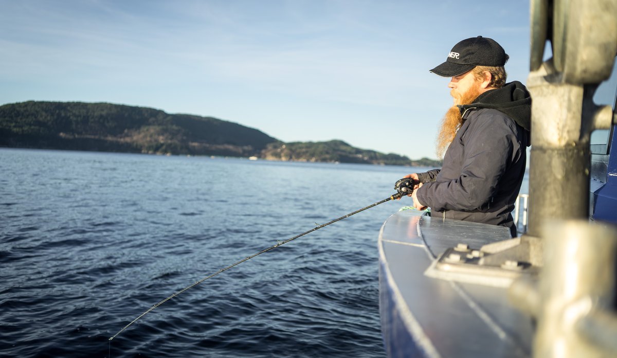 
Mann med blå caps og langt skjegg står i båt med fiskestand over rekka og fisker.