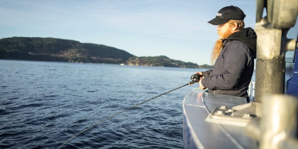 

Mann med blå caps og langt skjegg står i båt med fiskestand over rekka og fisker.