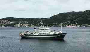 Forskningsskip på vei mot havn med berg og hus i bakgrunnen.