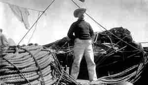 En mann med hatt står bredbent på dekk blant taljer og trosser,