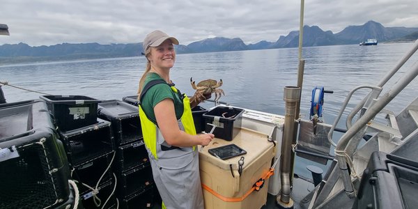 

Forsker på dekk med fiskebukse, måleutstyr og krabbe i hånda. Fjord og fjell i bakgrunnen