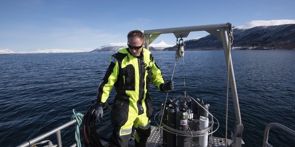 

Mann i kjeledress bak på lettbåt med en vannhengerkrans stående på dekk.