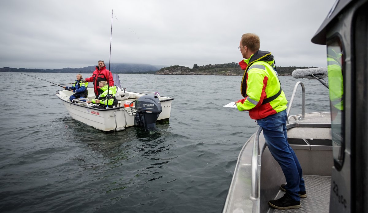 
Mann i en båt holder notatblokk i hendene mens han snakker med tre fiskere i en annen båt.