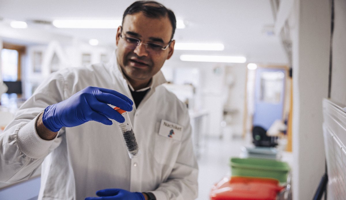 En mannlig forsker i hvit frakk er på et laboratorie. I hånden holder han et rør med en sedimentprøve