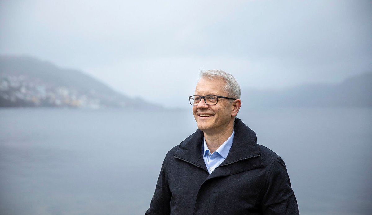 
Portrett av Nils Gunnar Kvamstø med lyseblå skjorte og mørk frakk. Hav, fjell og skyer i bakgrunnen.