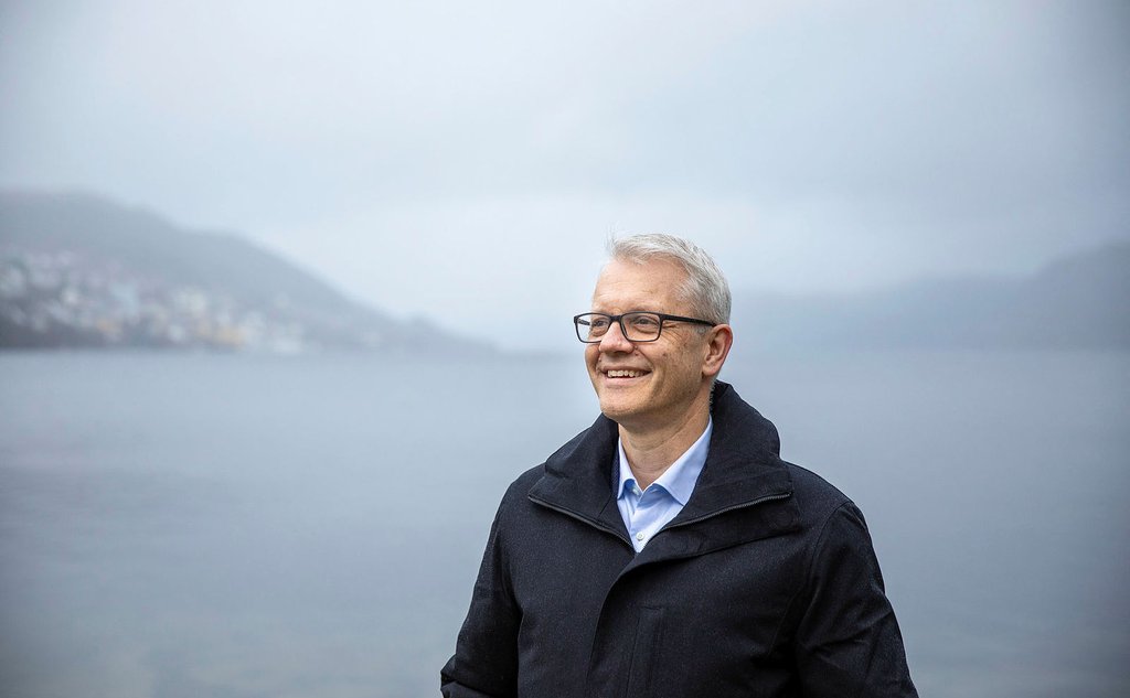 Portrett av Nils Gunnar Kvamstø med lyseblå skjorte og mørk frakk. Hav, fjell og skyer i bakgrunnen.