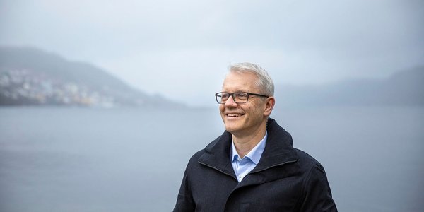 

Portrett av Nils Gunnar Kvamstø med lyseblå skjorte og mørk frakk. Hav, fjell og skyer i bakgrunnen.