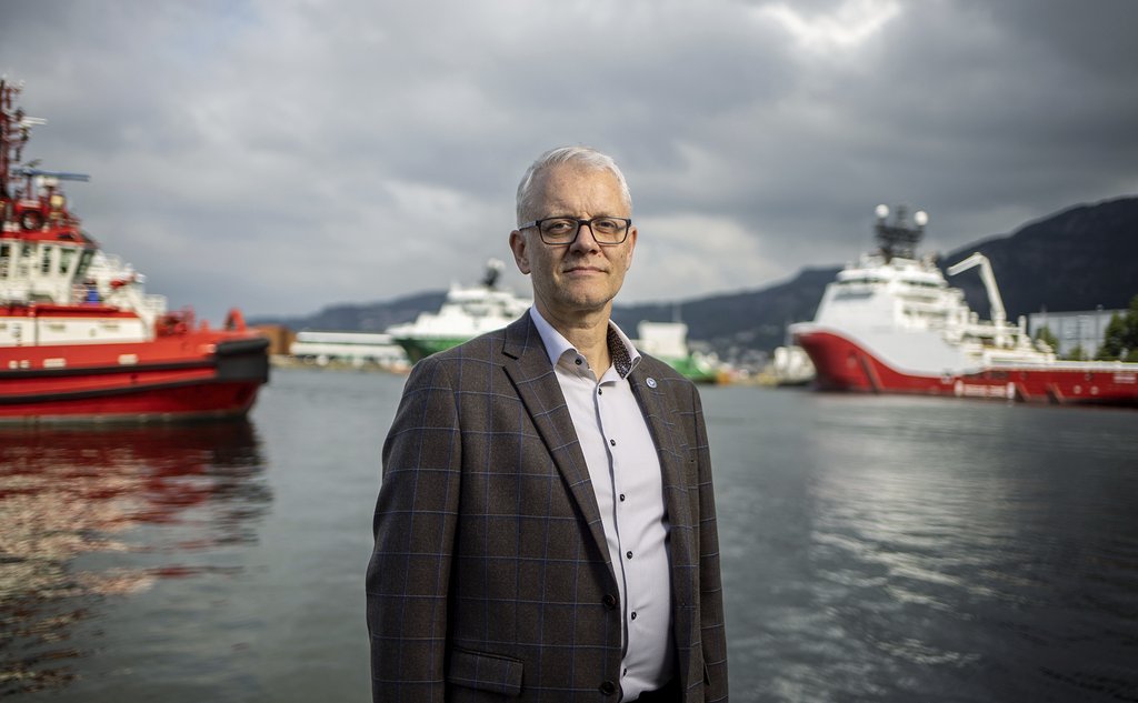 En mann med briller og i dressjakke står mot kameraet. i bakgrunnen skimter en havet og noen båter
