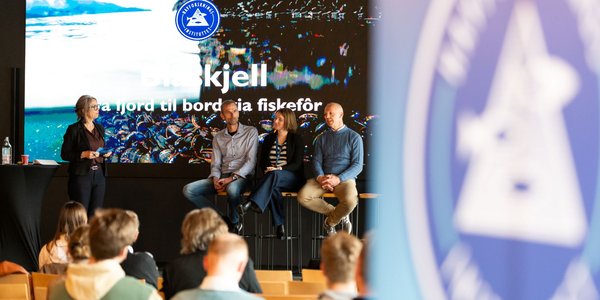 

Bilde fra et arragement. På scenen sitter tre paneldeltagere på hver sin stol. Ordstyrer står