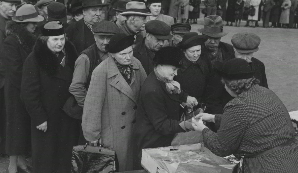 Svarthvittbilde av masse mennesker i kø, de fleste med kåpe eller frakk og hatt. En fiskehandler står med ryggen til oss. 