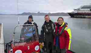 To menn og en kvinne, alle dykkere, står i en båt langs kaien. I bakgrunnen ser man så vidt Hurtigruta.