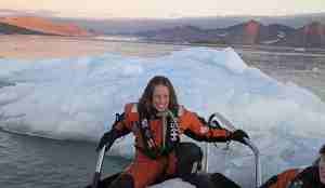 En kvinne sitter i bauen på en småbåt. Foran henne i båten ligger en stor isklump. Bak henne ser en isen ligge i fjorden. 