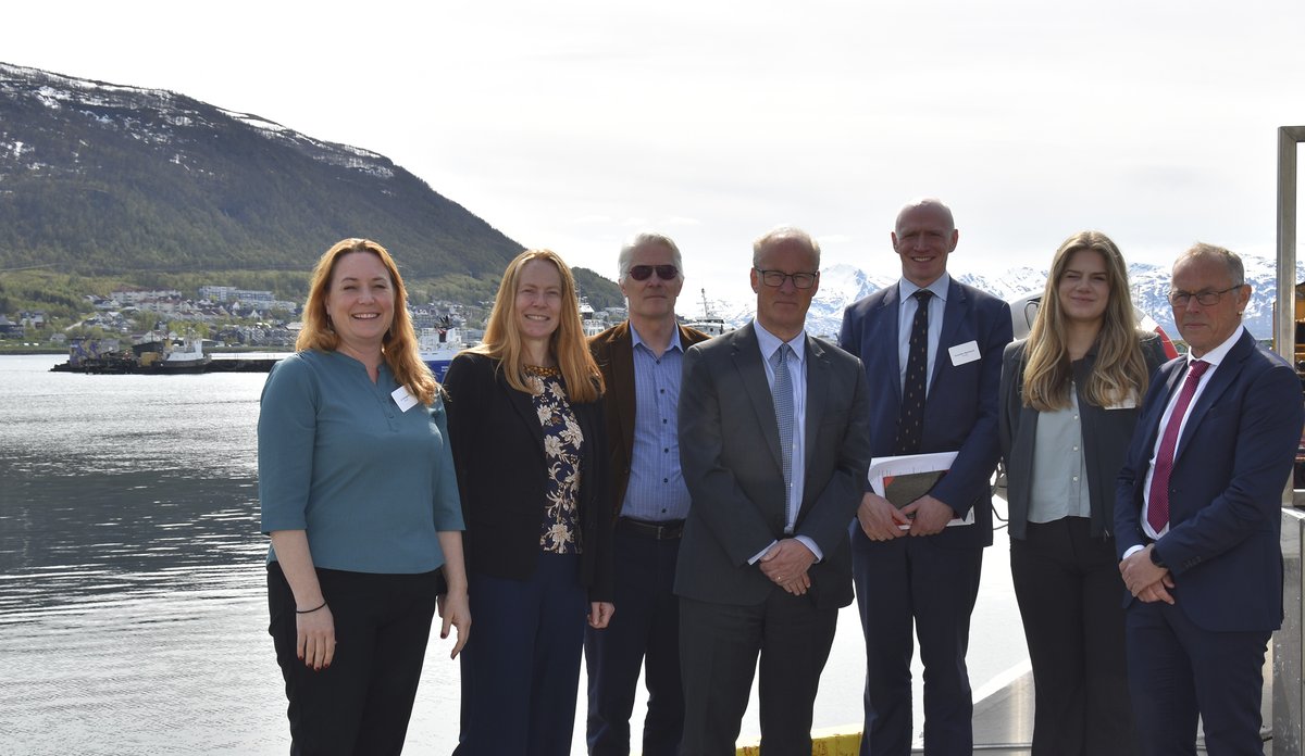 
Syv personer står oppstilt på en kai med utsikt mot havet bak seg. 