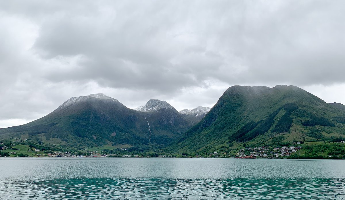 
Bygd som ligger nedenfor fjeller med en fjord i forgrunnen under grå skyer