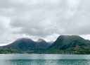 Bygd som ligger nedenfor fjeller med en fjord i forgrunnen under grå skyer