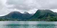 

Bygd som ligger nedenfor fjeller med en fjord i forgrunnen under grå skyer