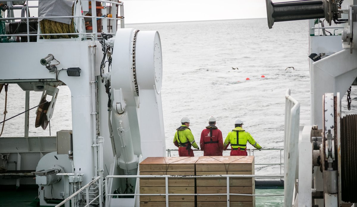 Tre personar i arbeidstøy på dekket til ein stor båt.