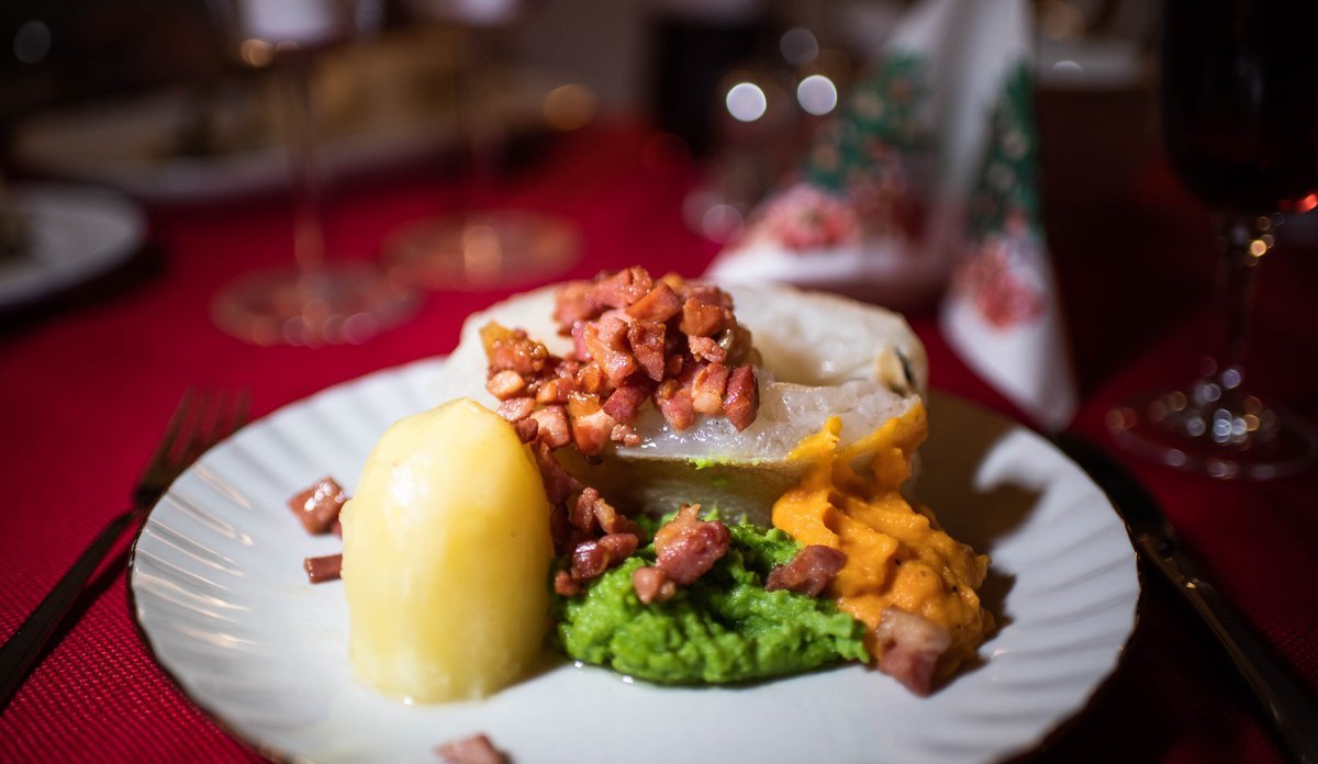 
Vi ser en hvit tallerken mot rød juleduk. På tallerkenen er det potet, ertepure, sprøstekt bacon og et stykke torsk.