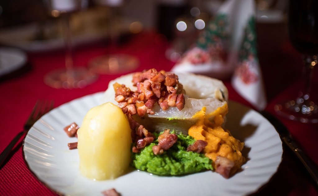 Vi ser en hvit tallerken mot rød juleduk. På tallerkenen er det potet, ertepure, sprøstekt bacon og et stykke torsk.