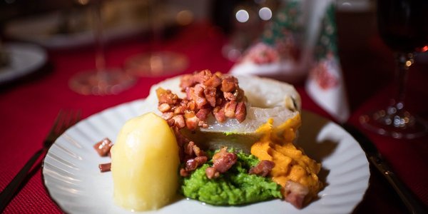 

Vi ser en hvit tallerken mot rød juleduk. På tallerkenen er det potet, ertepure, sprøstekt bacon og et stykke torsk.