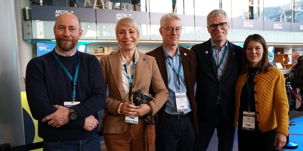 

Fem smilande personar oppstilte på rad i ein konferansehall.