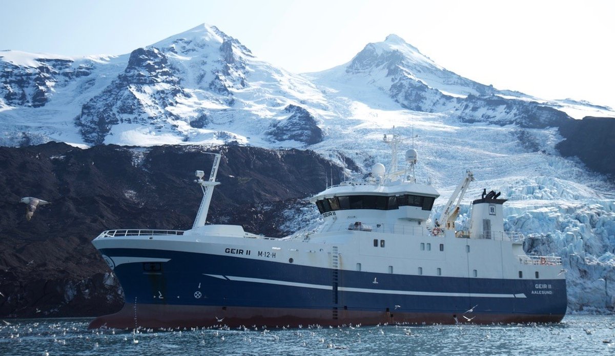 Blå og hvis fiskebåt foran øy med snøkledde fjell og en isbre.